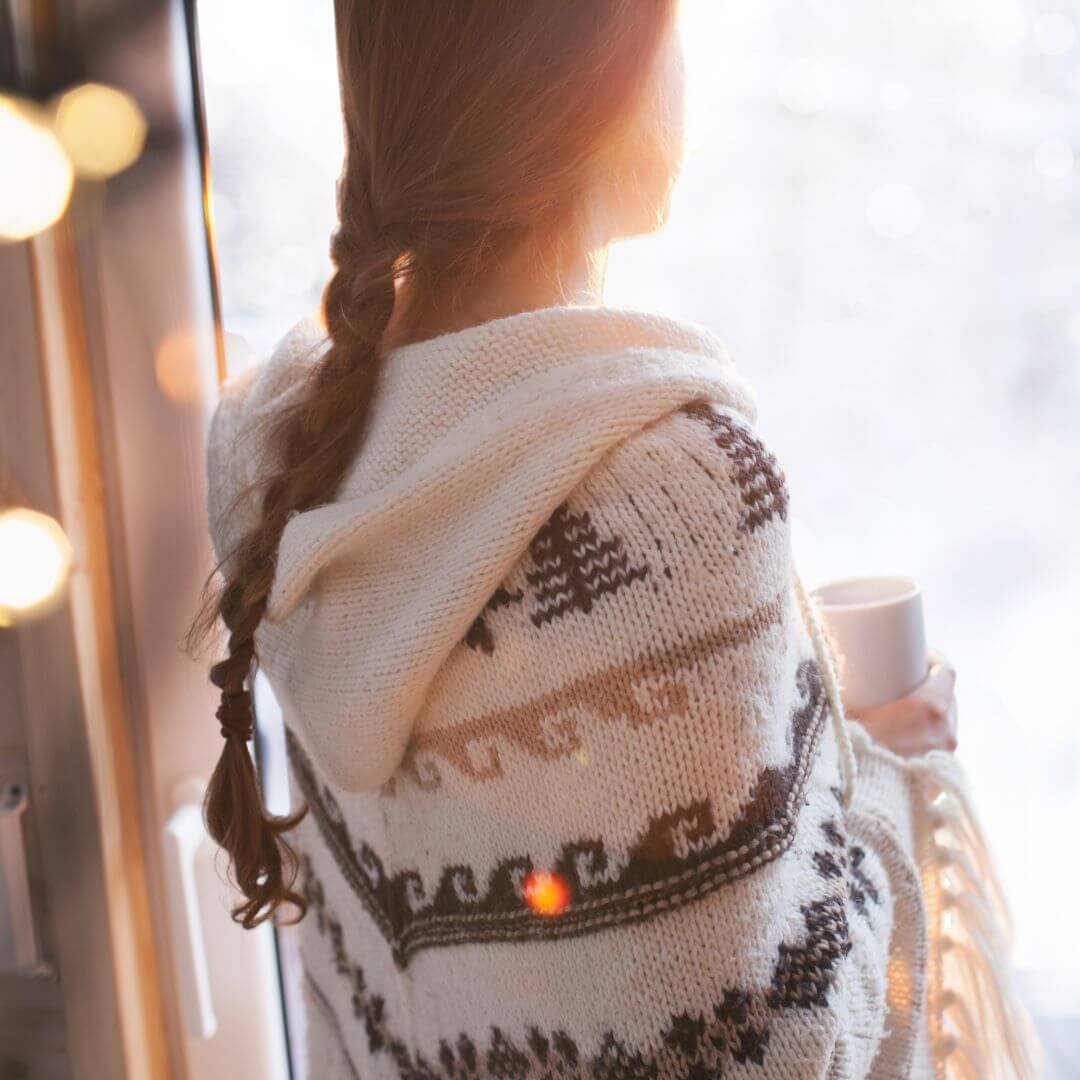 woman looking out window wearing warm jumper and hot collagen drink